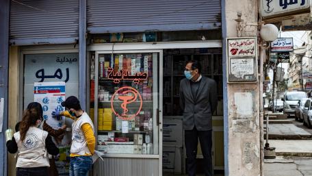 A man stands in front of a pharmacy while two volunteers hang a poster providing instructions on how to protect from COVID-19 